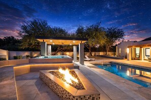 Un espace piscine extérieur moderne doté d’un jacuzzi, d’un foyer et d’un coin salon couvert, le tout sous un ciel étoilé.