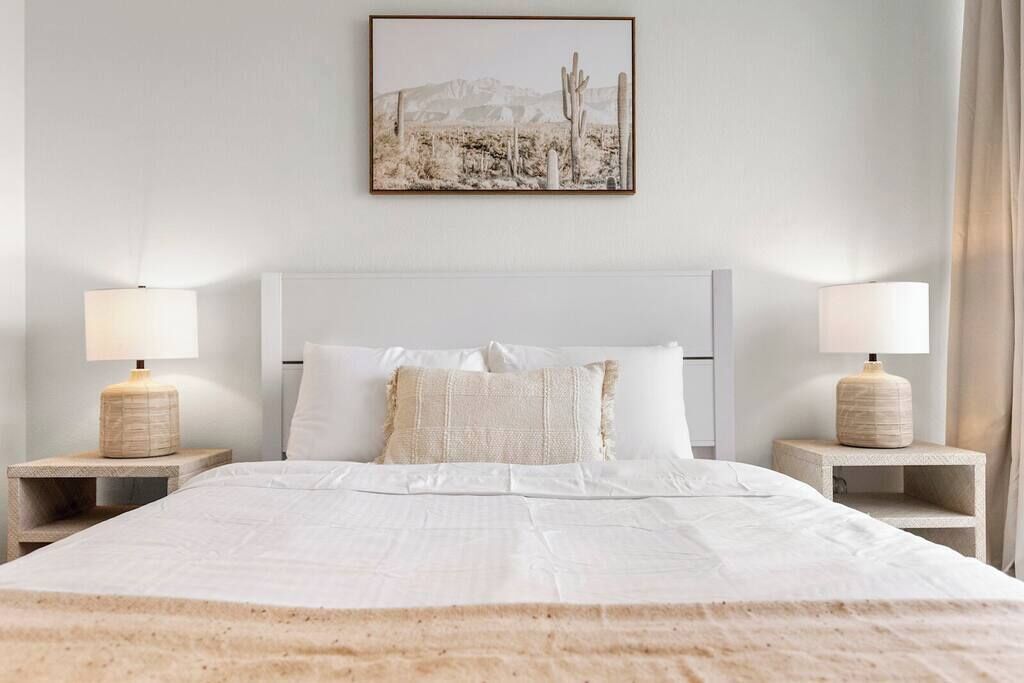 A neatly made bed with white linens, two bedside tables with lamps, and a framed picture of a desert landscape on the wall.