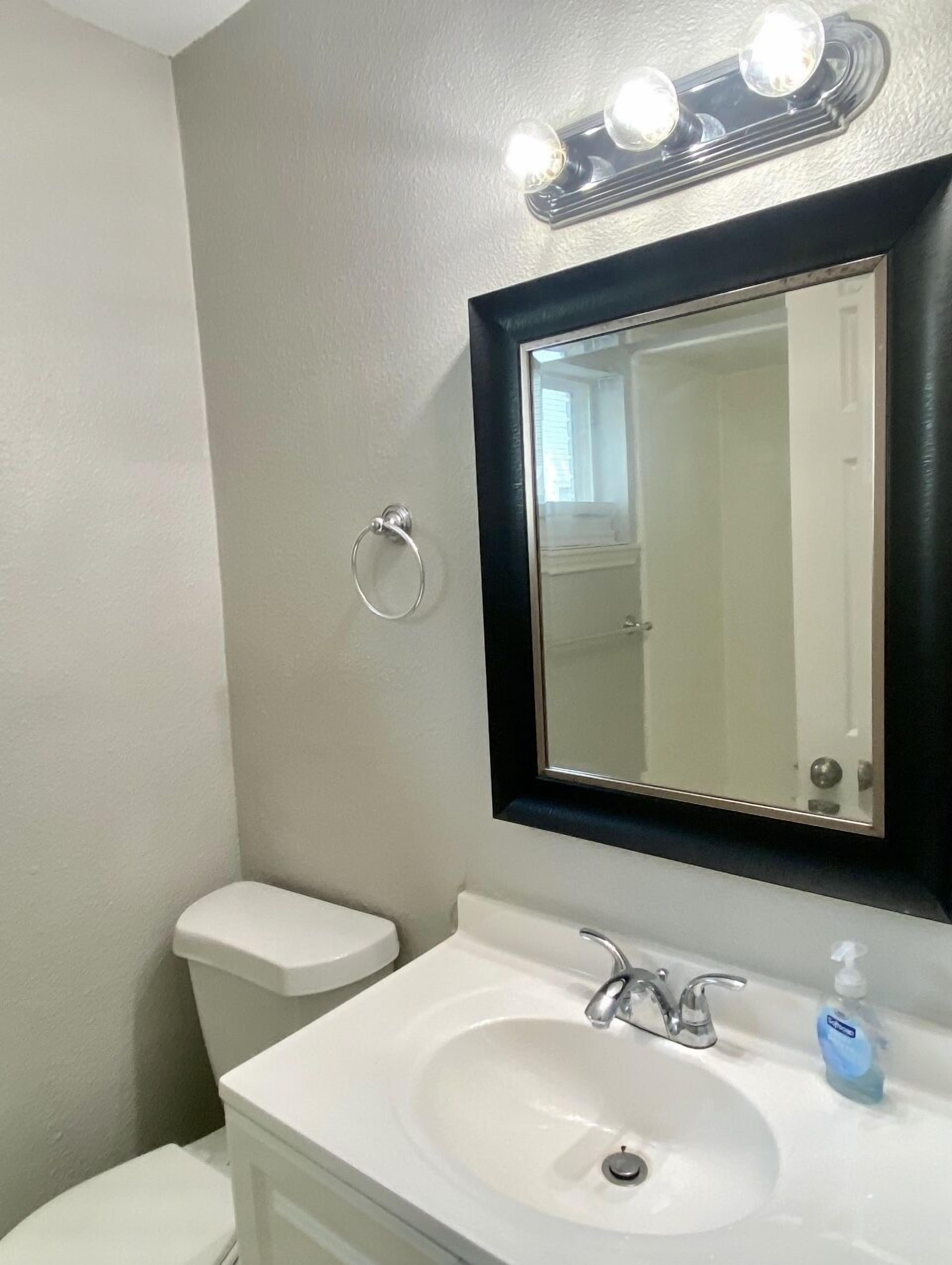 A bathroom with a white sink, a mirror, and a toilet. There are three lights above the mirror.