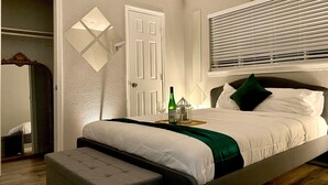 Ein ordentlich bezogenes Bett mit einer grünen Decke, ein Nachttisch mit Flasche und Glas sowie ein Fenster mit Jalousien.