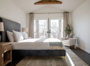A modern bedroom with a large bed, bedside tables, a chair, and a view of the outdoors through a glass door. - Stone Ridge Modern Mountain House (Stone Ridge)