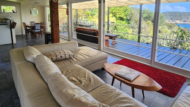 Ampla sala de estar com um sofá bege grande, mesa de centro de madeira e tapete vermelho. Uma área de jantar com mesa e cadeiras é visível através de uma porta de vidro que dá para um deck externo com vista para vegetação e um corpo d'água.
