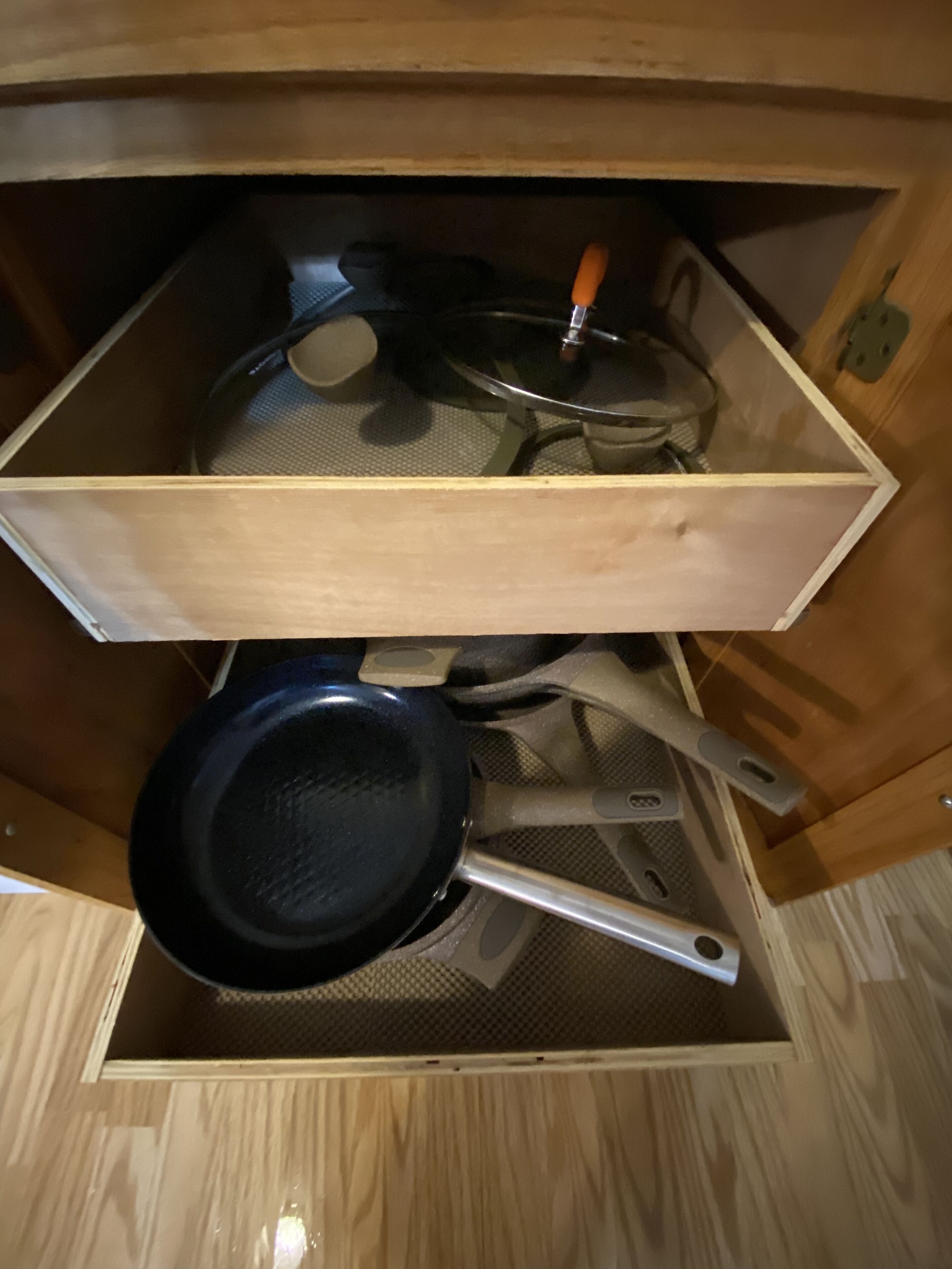 A wooden drawer with a metal mesh interior, containing a frying pan, a spatula, and a pot.