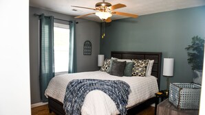 Une chambre à coucher avec un lit, un ventilateur de plafond, une fenêtre avec des rideaux, une lampe de chevet et une plante.
