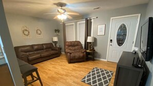 Un salon avec un canapé en cuir marron, un fauteuil inclinable, un plancher en bois, un ventilateur de plafond, une porte avec une fenêtre et un téléviseur posé sur un meuble.