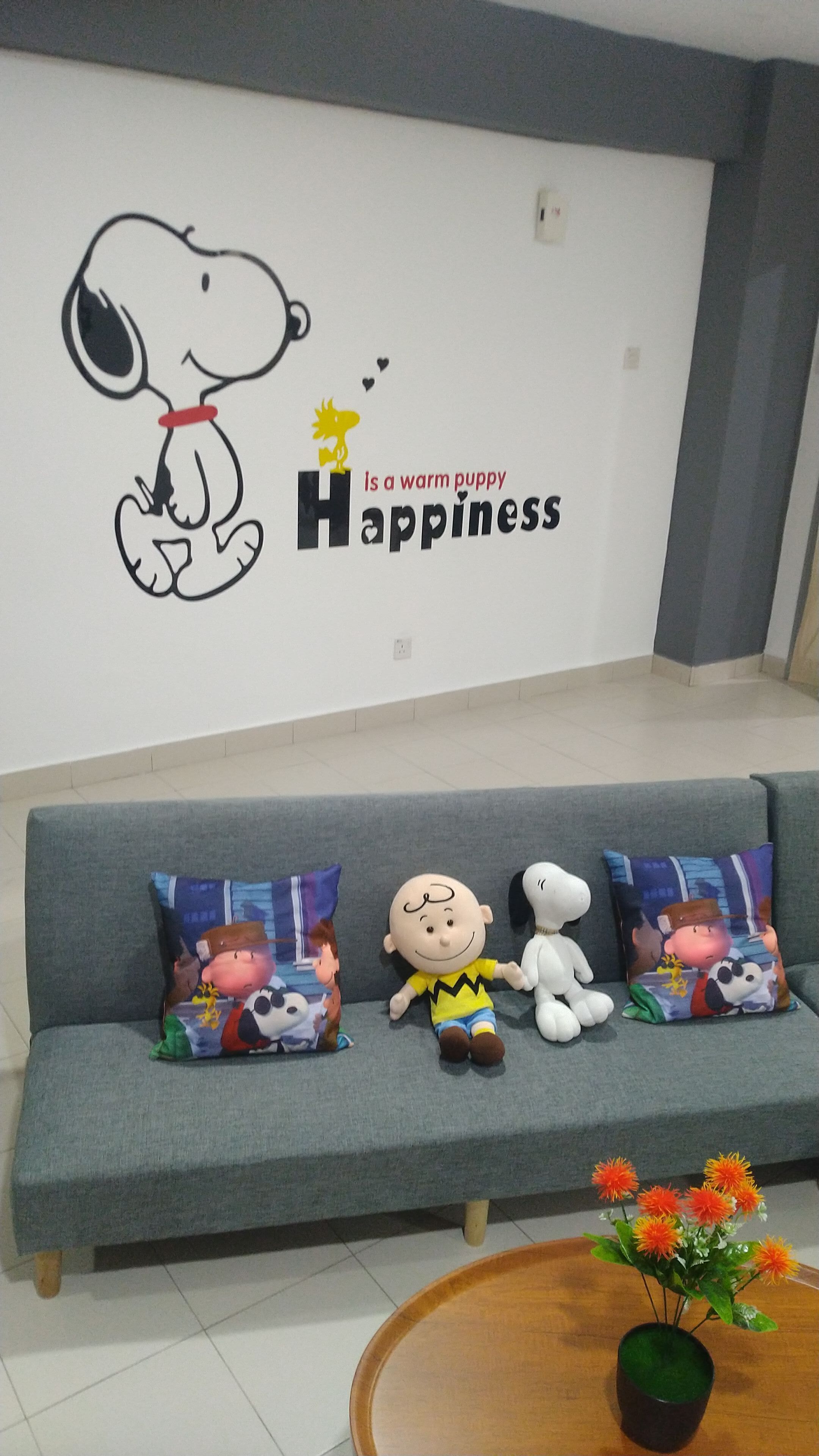 A living room with a wall featuring a Snoopy mural and the text "Happiness is a warm puppy". There is a grey sofa with three Snoopy-themed pillows and a small table with a flower vase.
