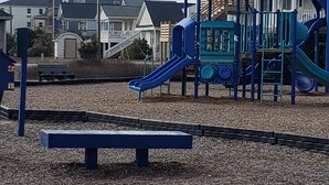 Un parque infantil con un tobogán azul y un pequeño refugio, rodeado de una zona de grava y edificios residenciales al fondo.