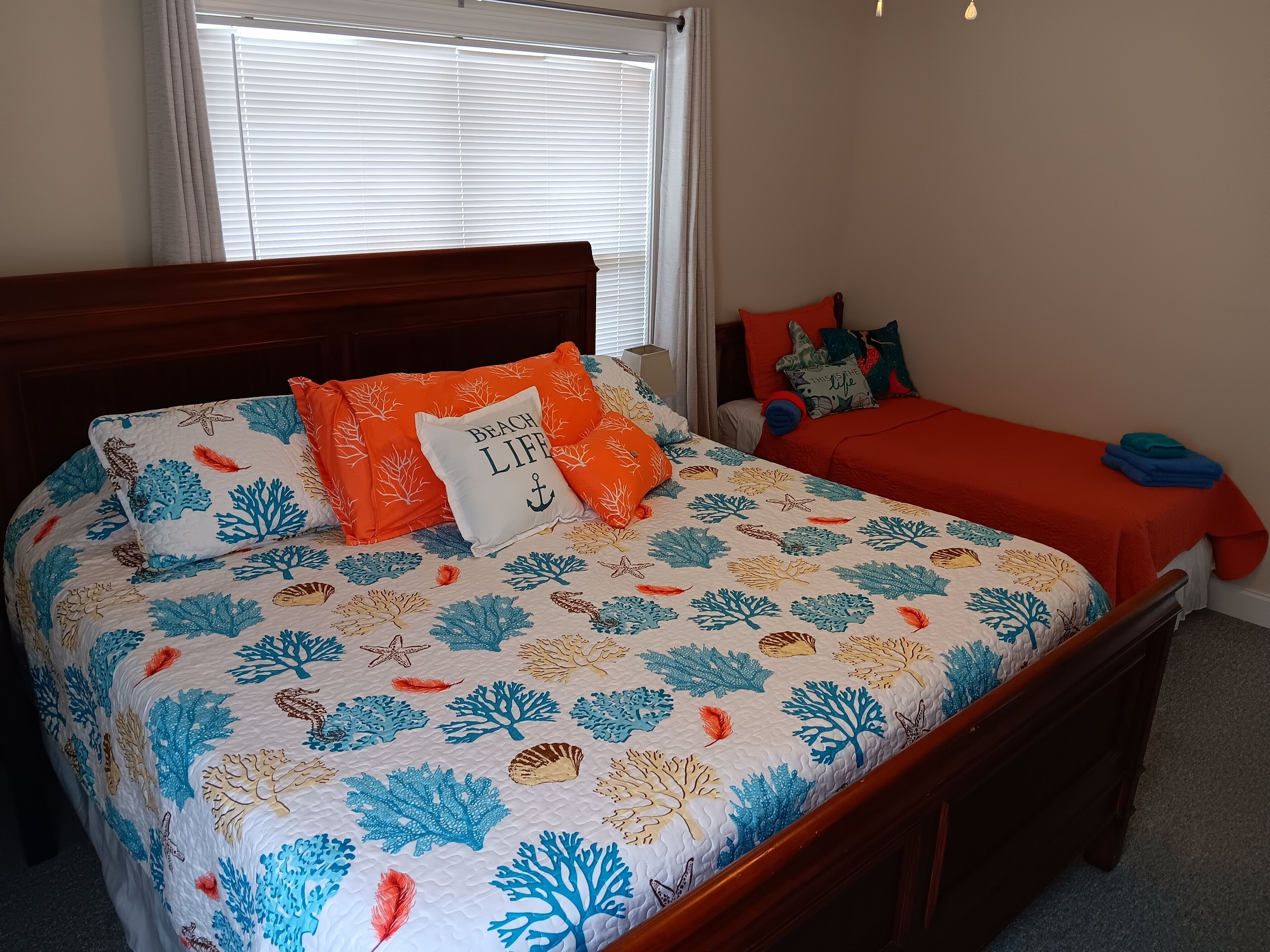 Un dormitorio con una cama que presenta un edredón con motivos de coral y vida marina, un cabecero de madera y una ventana con persianas.