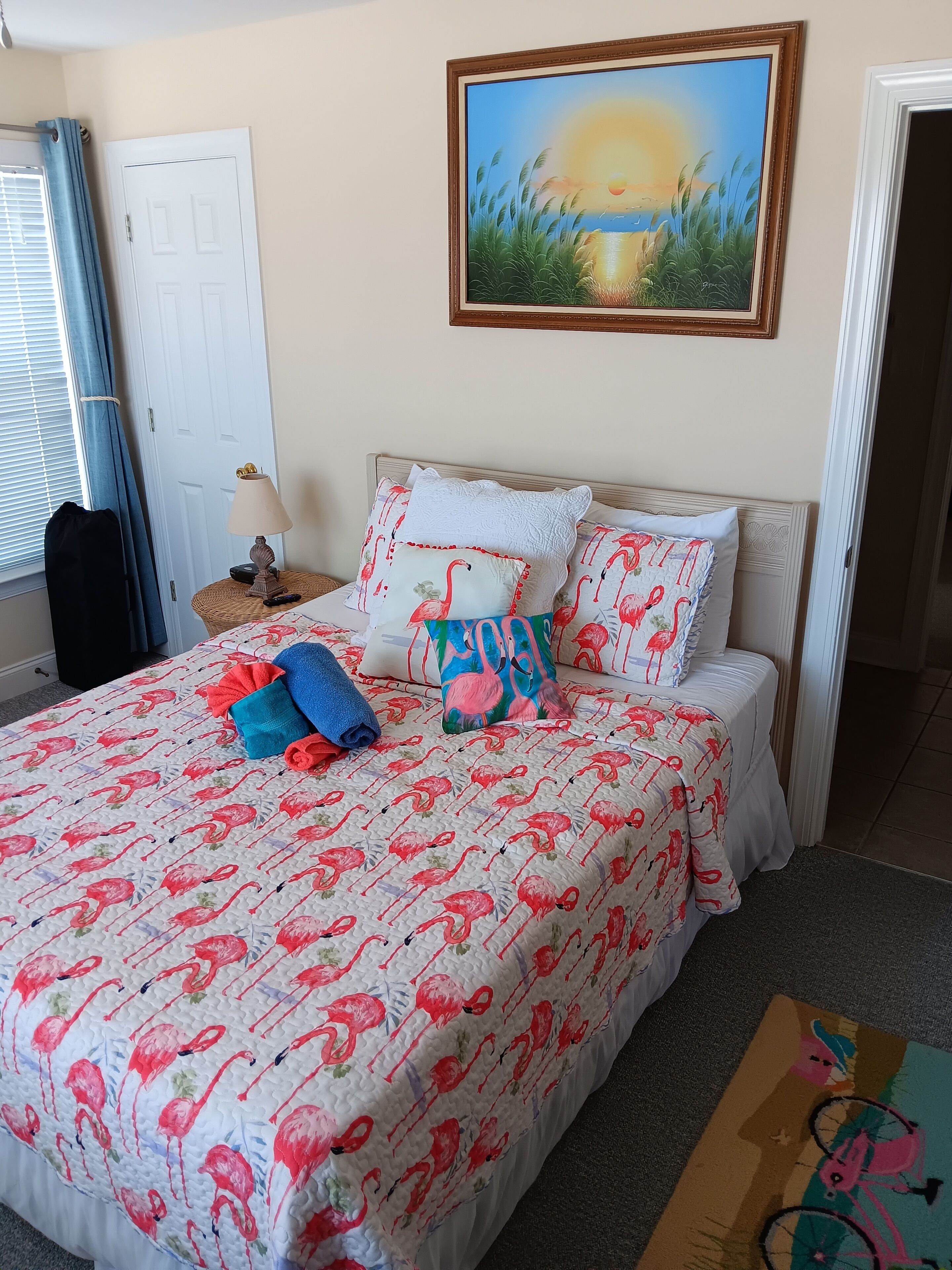 A bedroom with a bed featuring a flamingo-themed duvet, a painting of a sunset, and a door with a curtain.