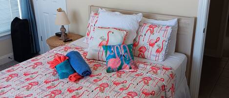 A bedroom with a bed featuring a flamingo-themed duvet, a painting of a sunset, and a door with a curtain.