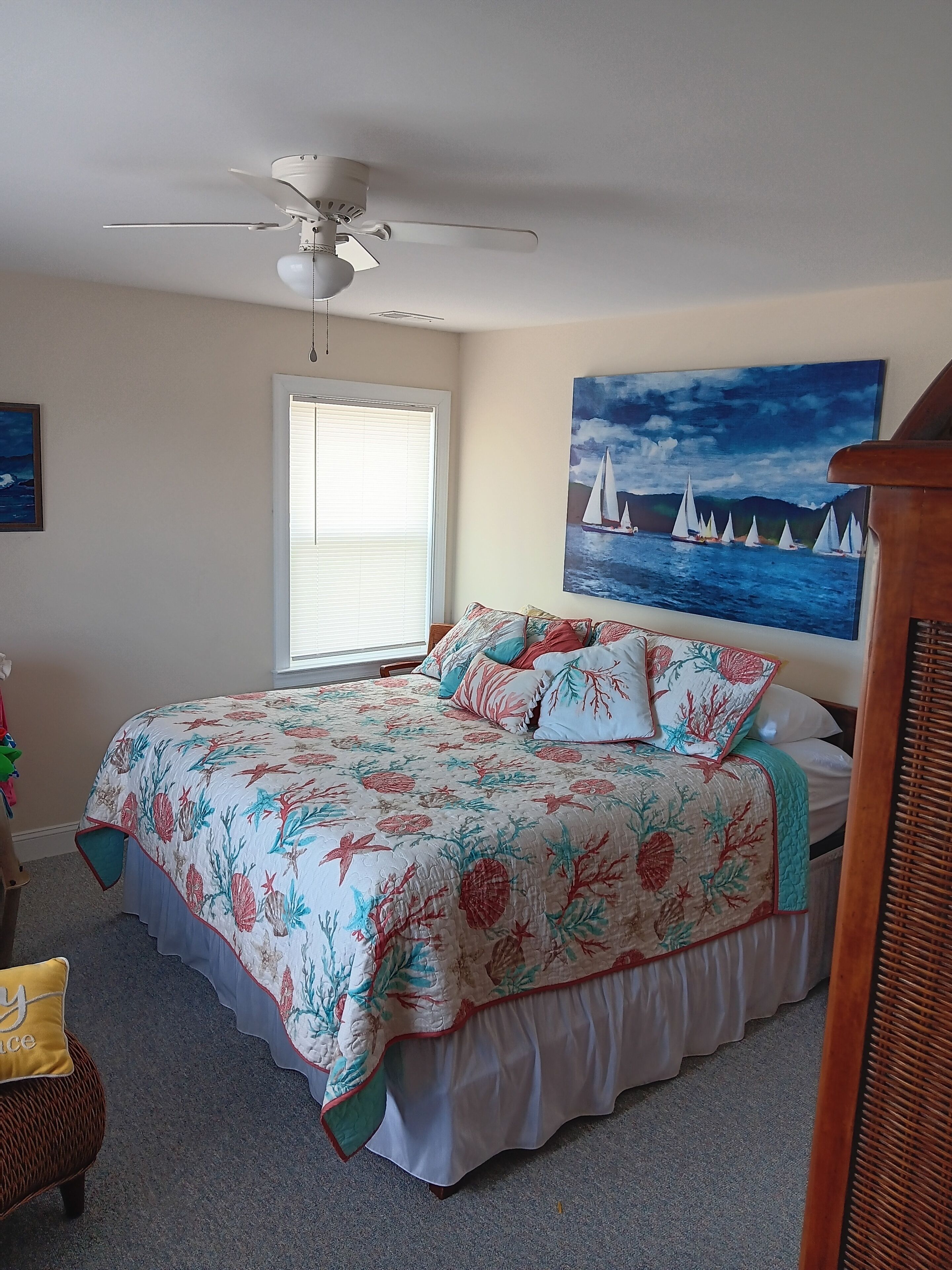 A bedroom with a bed, a ceiling fan, a window with blinds, and a painting of sailboats on the wall.