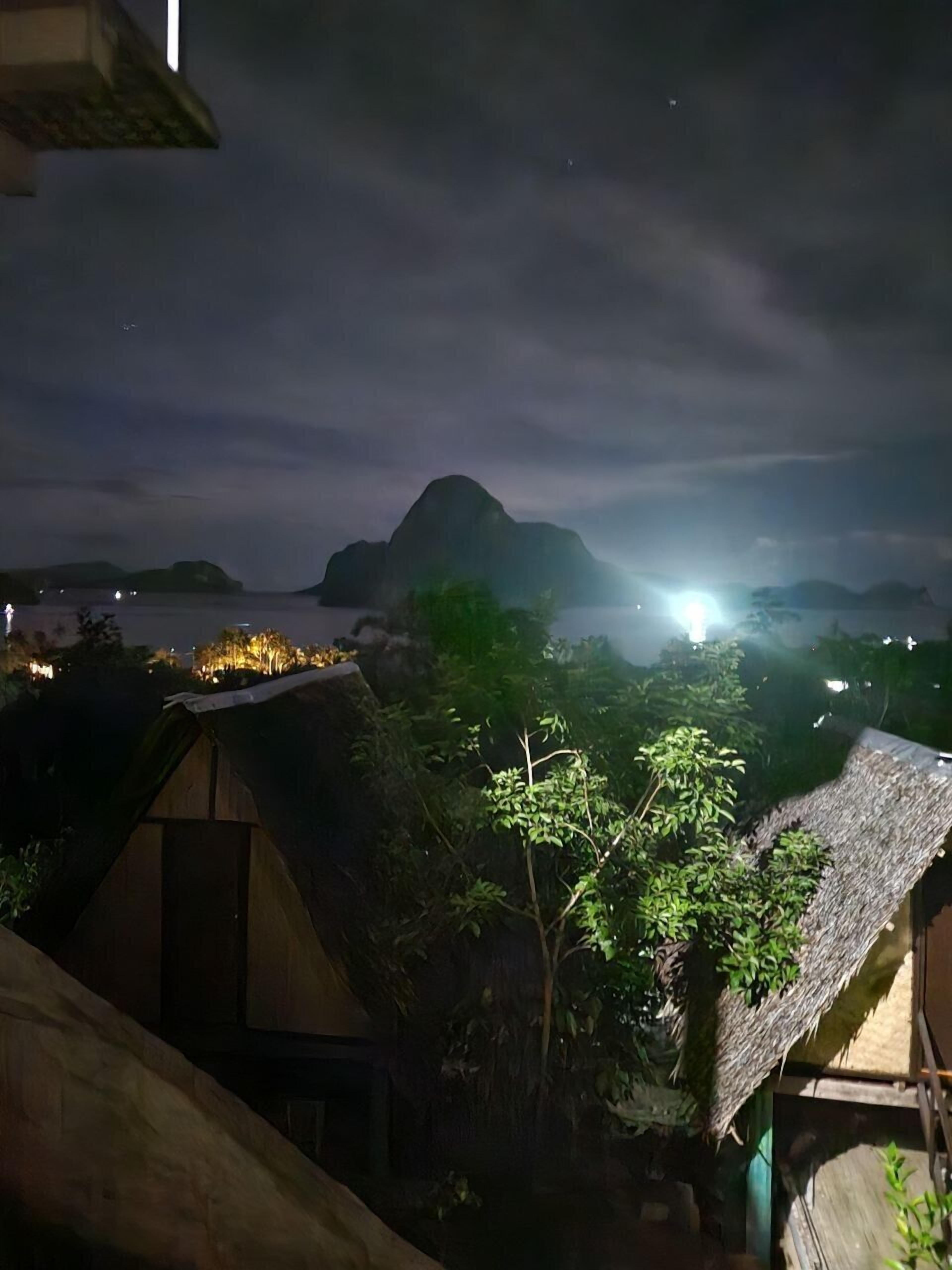 Vista nocturna de una zona costera con edificios de techo de paja, una montaña al fondo y una fuente de luz brillante.