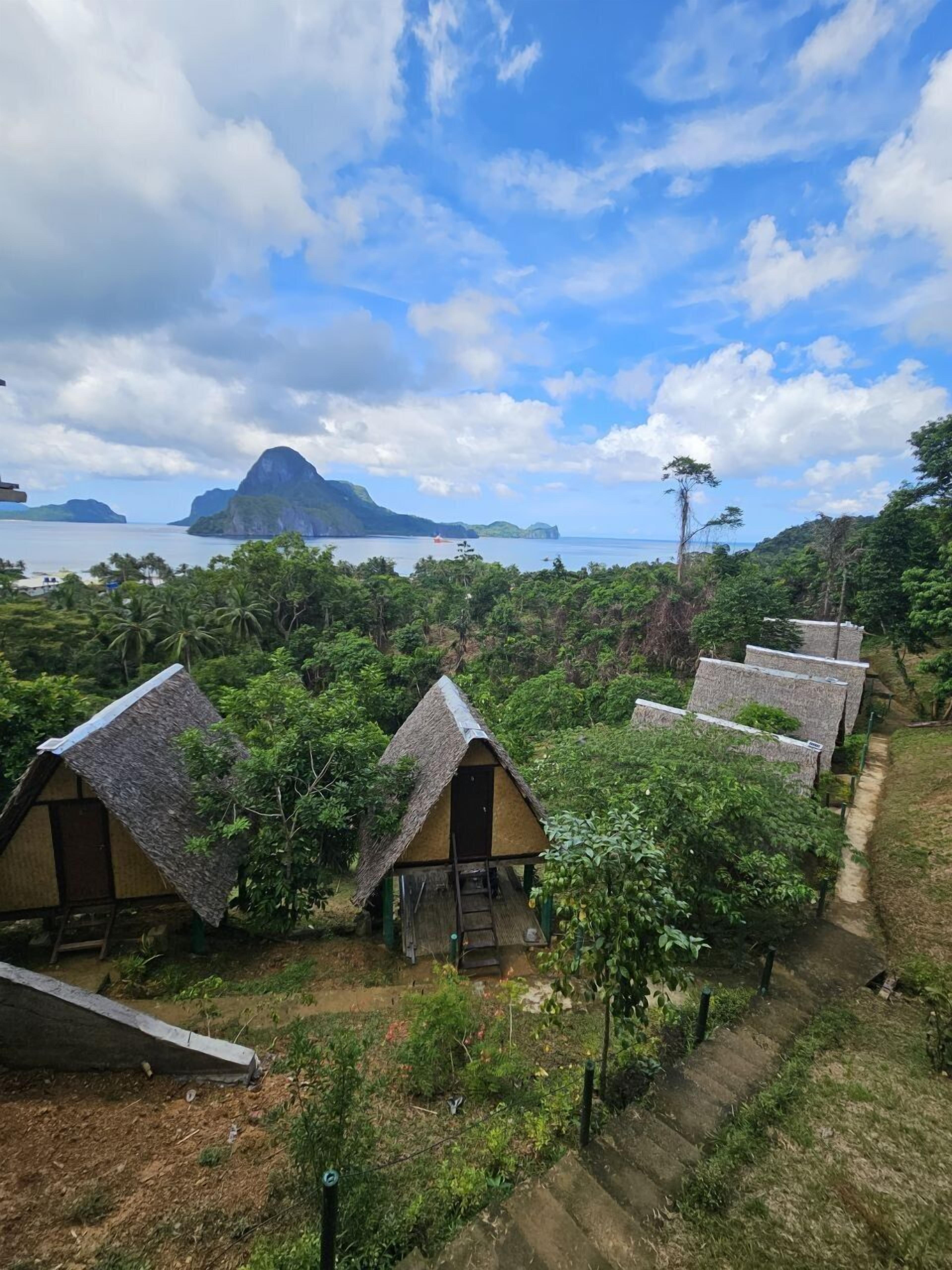 A row of thatched-roof cottages with a staircase leading up to a wooden deck, surrounded by lush greenery and overlooking a body of water with distant islands.