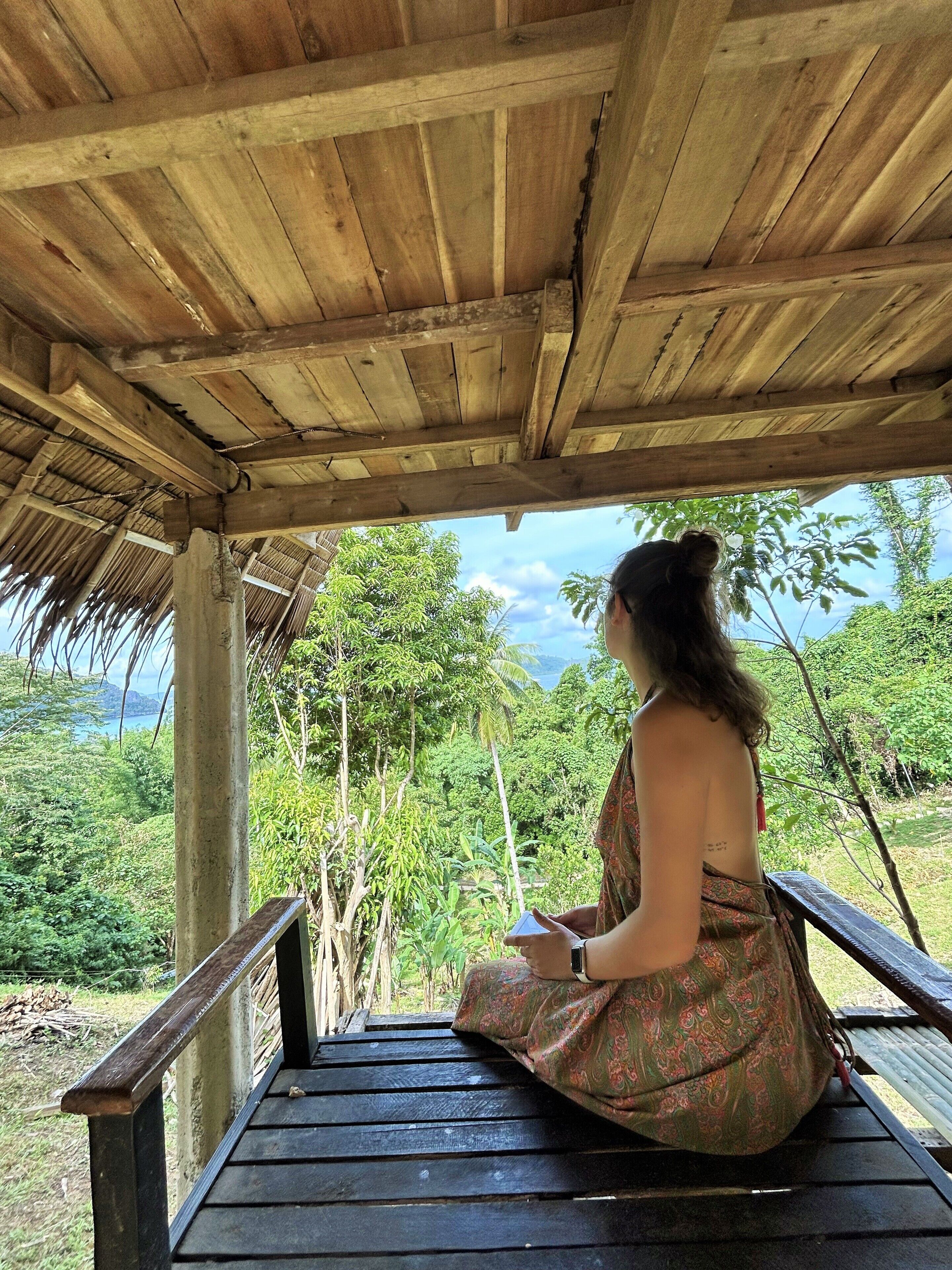 A person sitting on a wooden bench under a wooden structure, overlooking a lush green landscape with trees and a view of the sea.