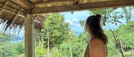 A person sitting on a wooden bench under a wooden structure, overlooking a lush green landscape with trees and a view of the sea.