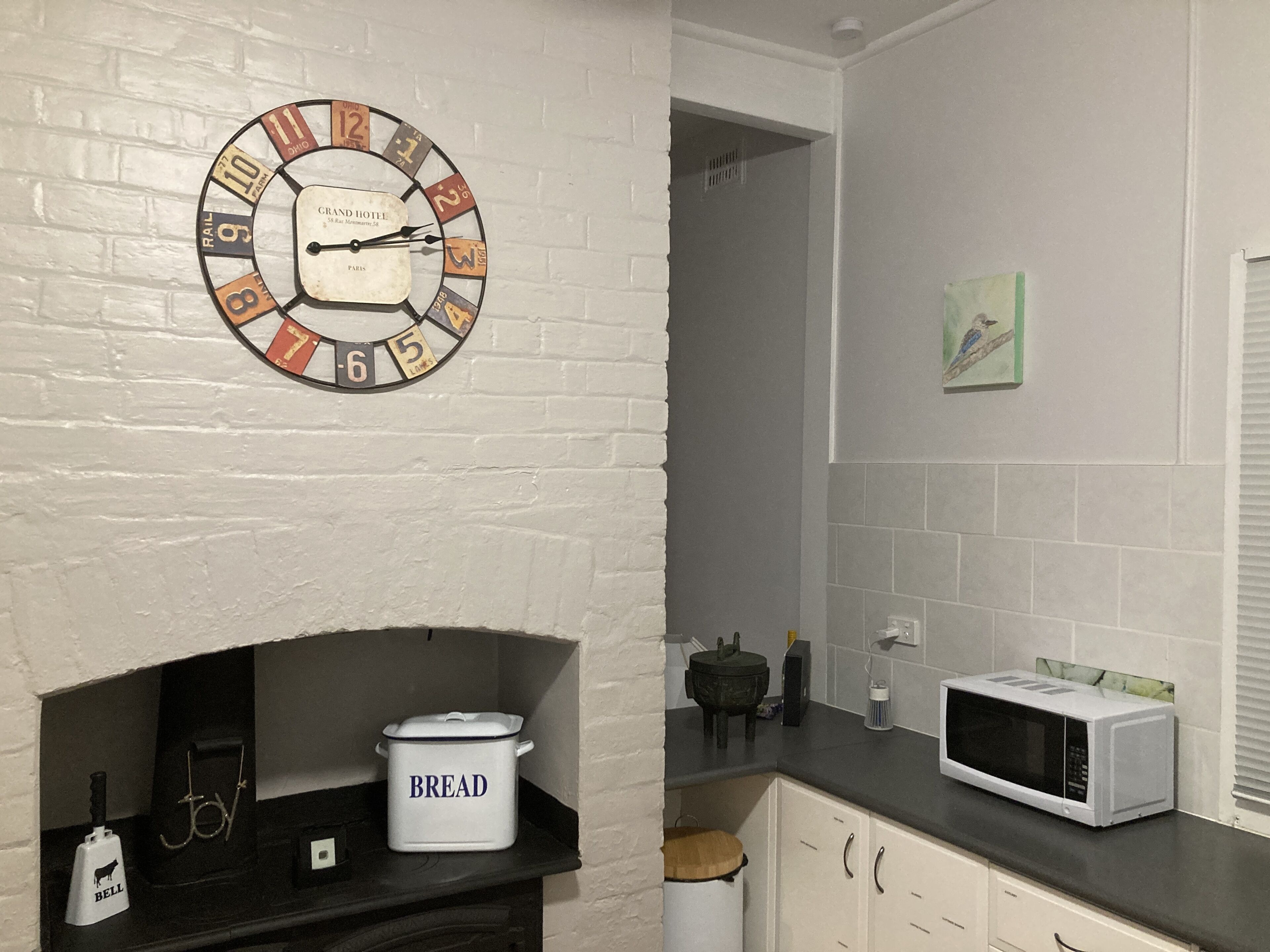 A kitchen with a white brick wall, a clock with numbers and letters, a bread container, a microwave, and a small cabinet.