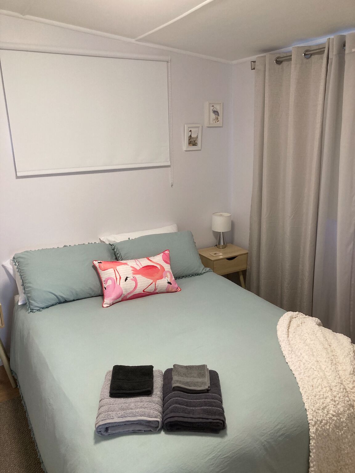 Un lit bien fait, recouvert d’une couvre-lit bleu clair, avec un coussin à l’effigie d’un flamant rose et deux serviettes pliées. Une table de chevet avec une lampe se trouve à côté du lit, et un petit cadre photo est accroché au mur.