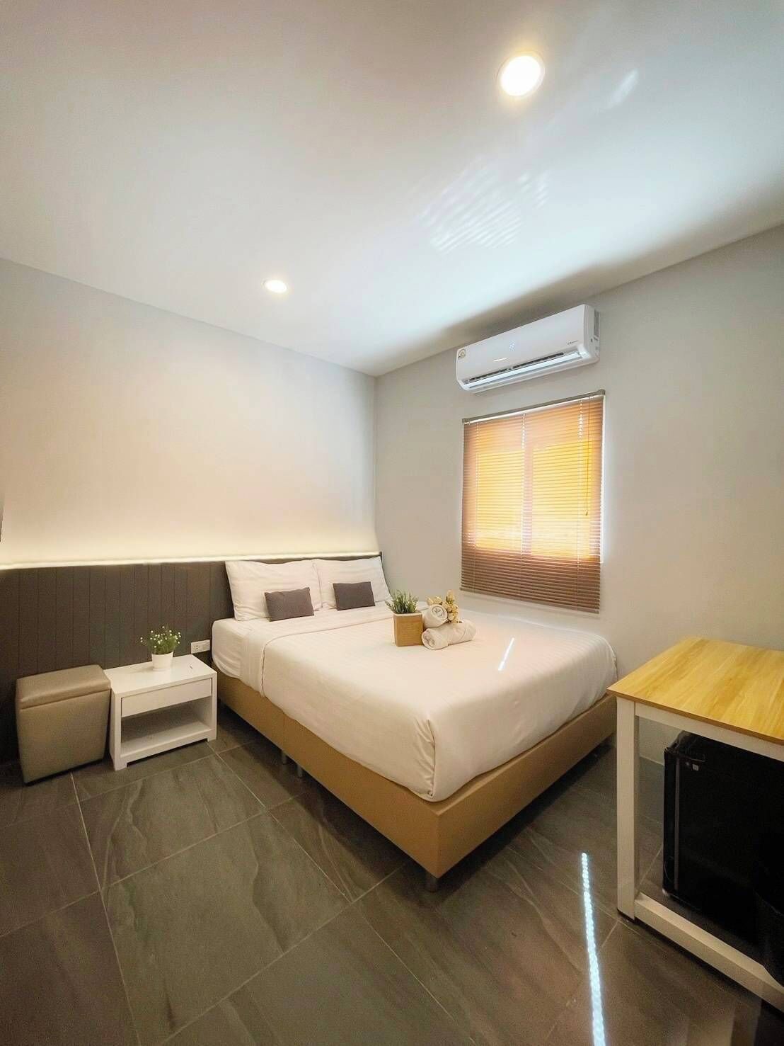 Een moderne hotelkamer met een groot bed, een nachtkastje, een klein nachtkastje, een airconditioningapparaat, een raam met zonwering en een houten tafel.