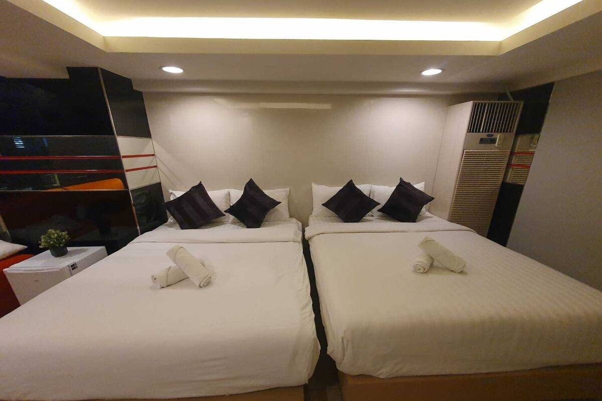 Une chambre d’hôtel avec deux lits, chacun doté de draps blancs et d’oreillers foncés, une petite table de chevet avec une plante et un système de climatisation intégré.