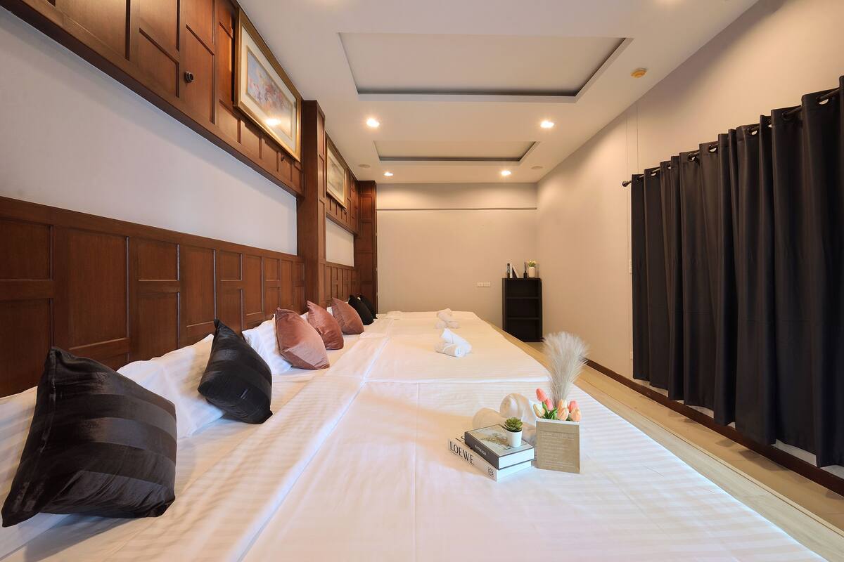 Une chambre d’hôtel avec deux lits, une tête de lit en bois, du linge de lit blanc, une petite table de chevet avec un livre et un vase de fleurs, et un rideau foncé à la fenêtre.