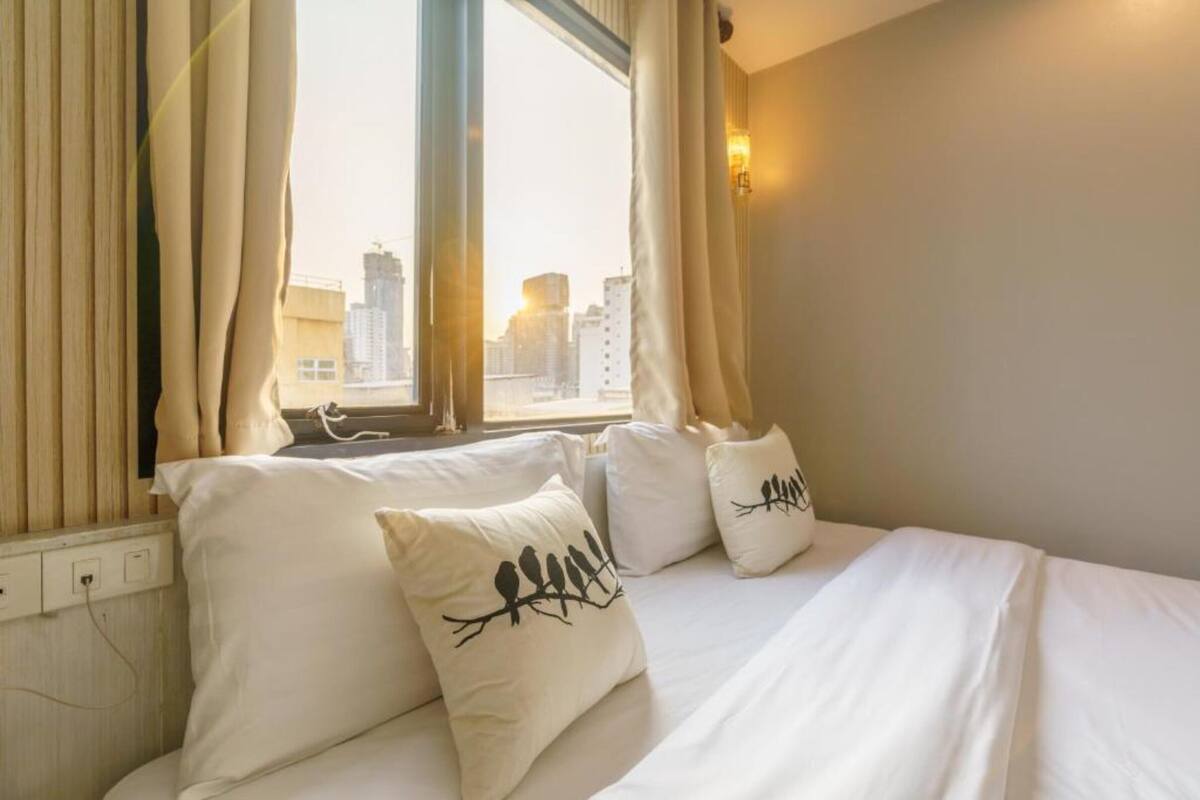 Une chambre d’hôtel avec un lit, deux oreillers ornés de motifs d’oiseaux, une fenêtre avec des rideaux et une vue sur la ville.