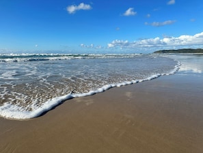Uma praia com ondas batendo na costa, céu claro e terra distante.