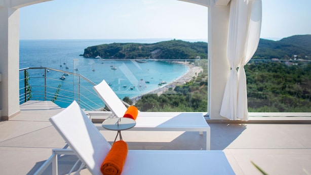 A balcony with a view of a beach and the sea, white furniture, and a glass railing.