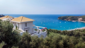 A coastal property with a white building, a balcony with pink flowers, and a view of the sea with boats.