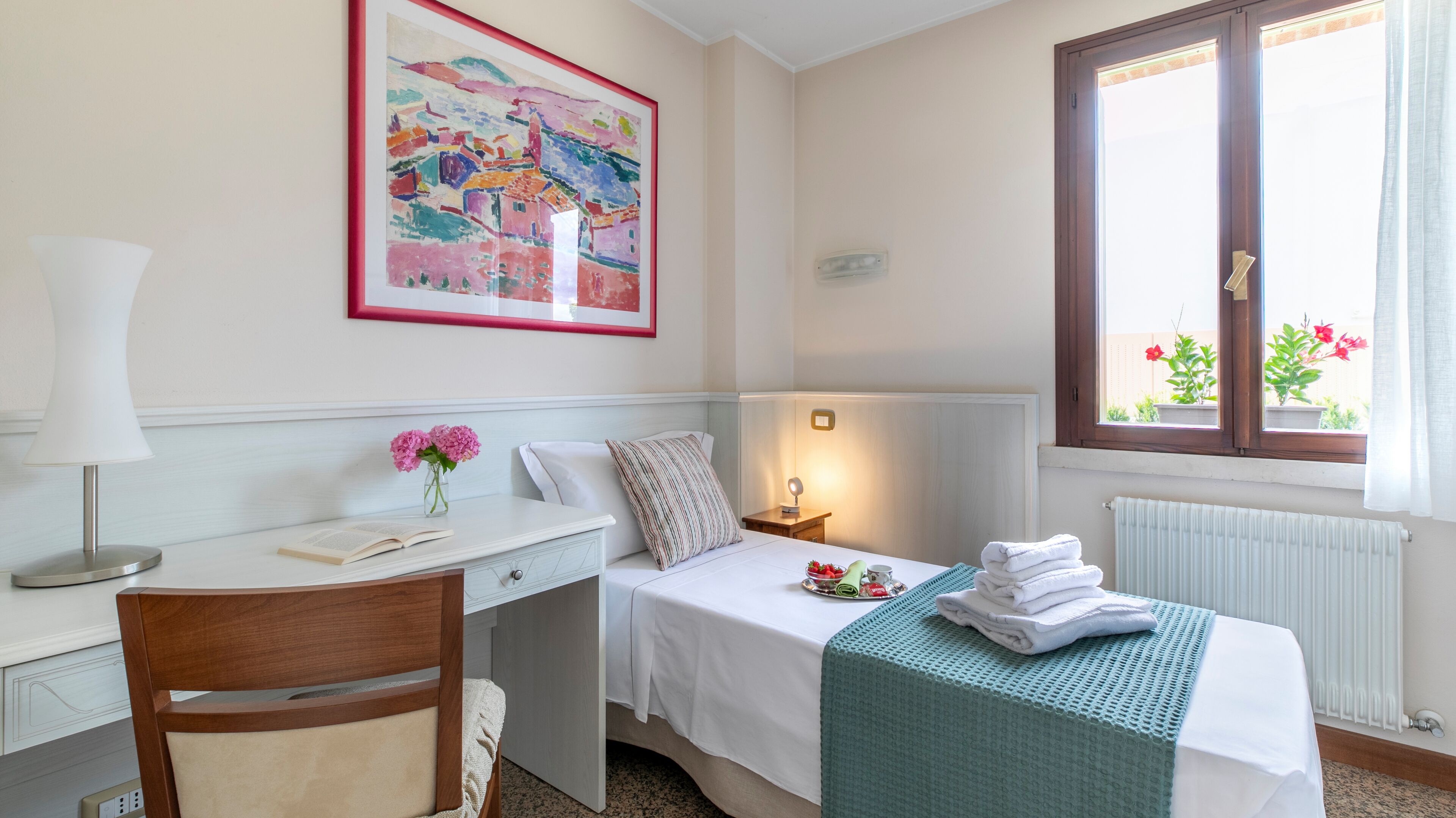 Ein Hotelzimmer mit einem Bett, einem Schreibtisch mit Lampe, einem Sessel, einem Fenster mit Blick auf Pflanzen und einem Bild an der Wand.