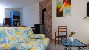 A living room with a patterned sofa, a wooden chair, a small table with a vase, a fireplace, and a kitchen area in the background. - Quiet and spacious cottage in the countryside (Fourilles)