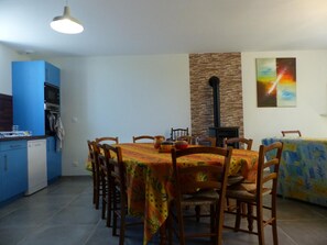 Een eetruimte met een ronde tafel, houten stoelen en een tafelkleed met een patroon. Links is een keuken met blauwe kastjes en een fornuis te zien.