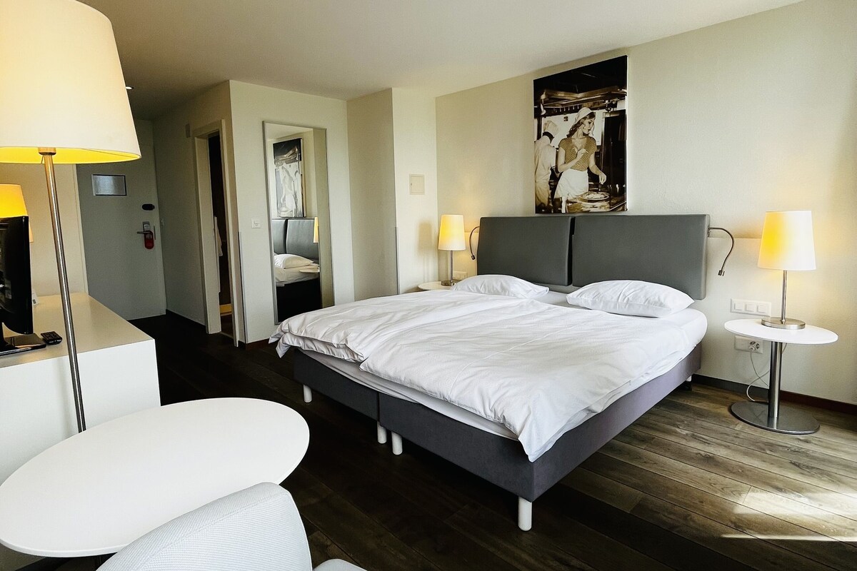 A hotel room with a bed, two bedside lamps, a chair, a small table, and a painting on the wall.