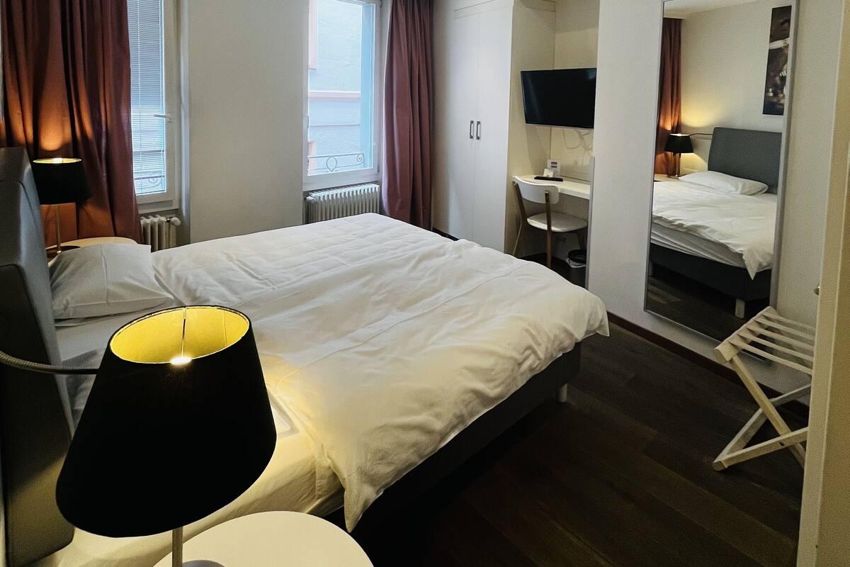 A hotel room with a bed, a desk, a chair, a lamp, a mirror, and a window with curtains.