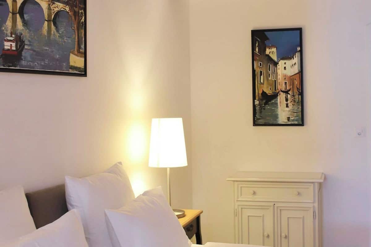 A hotel room with a large bed, a nightstand with a lamp, and two paintings on the walls.