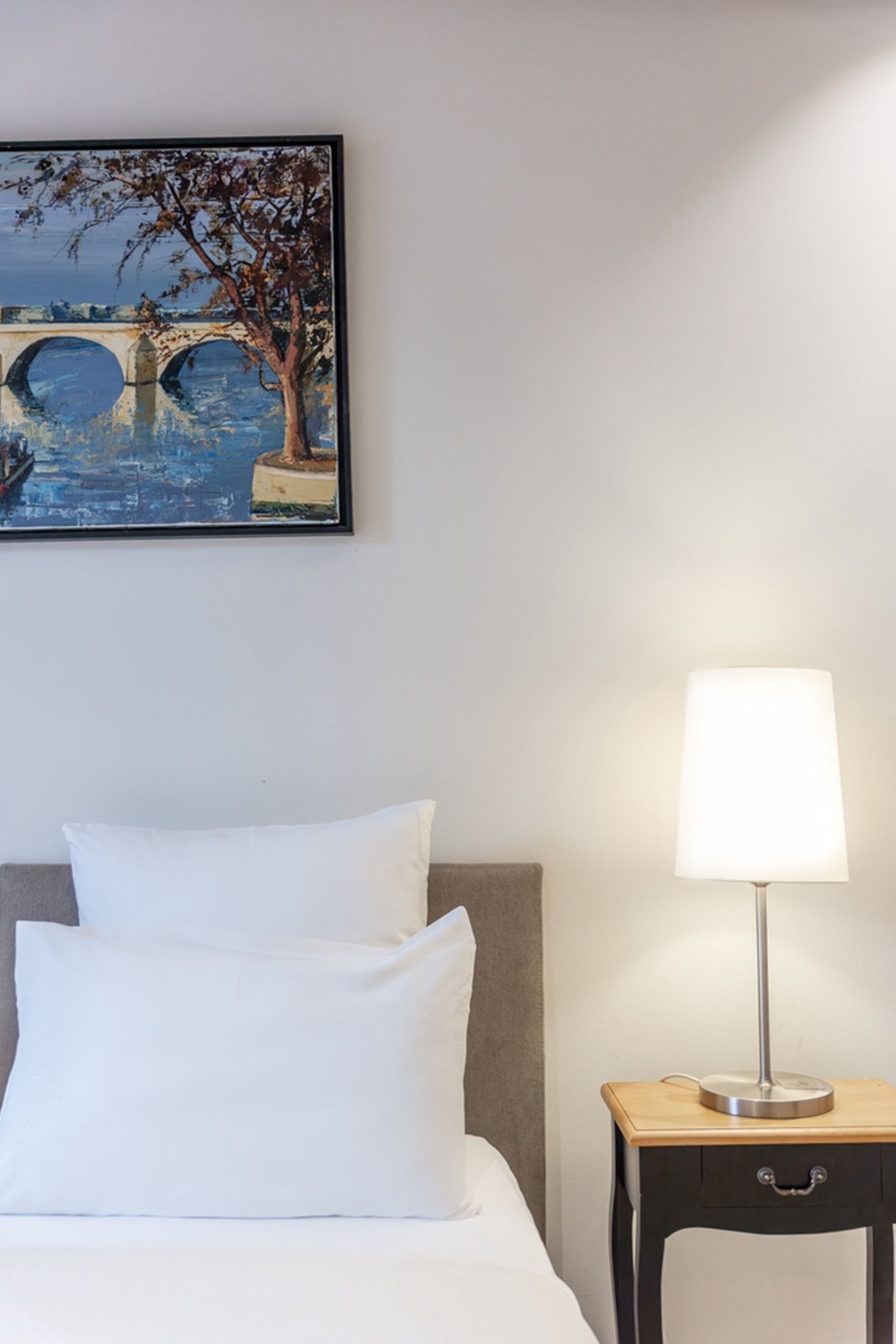 Una cama bien hecha con sábanas blancas, una lámpara de noche y un cuadro enmarcado de un puente y un árbol en la pared.