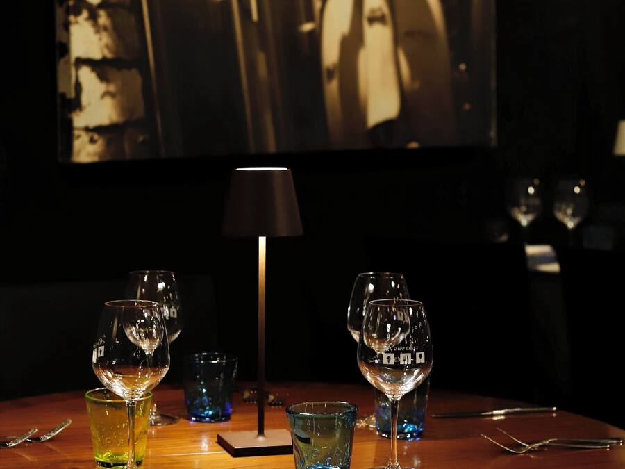A dimly lit room with a table set for dining, featuring two wine glasses and a small blue glass. A lamp is positioned in the center, and a large framed picture hangs on the wall behind the table.