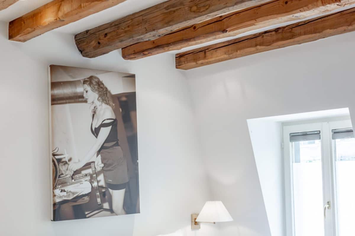 A bedroom with a wooden ceiling, a bed with white bedding, a wall-mounted lamp, and a window.