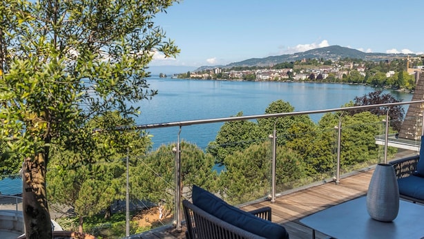 Eine Terrasse mit Blick auf einen See, umgeben von Bäumen und einer entfernten Stadtlandschaft.