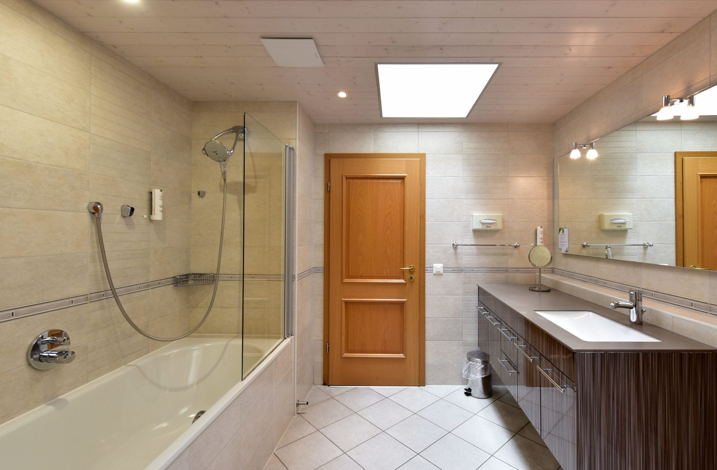 A modern bathroom with a glass-enclosed shower, a wooden door, a vanity with a mirror, and a large window above the sink.
