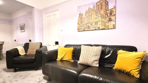 A living room with a black leather sofa and armchair, a dining table with chairs, and a painting of a cathedral on the wall. - 2 Bed Holiday Home with on-street parking and river walks (York)