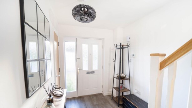 A hallway with a wooden floor, a white door, a black metal shelf with a bowl, and a hanging light fixture.