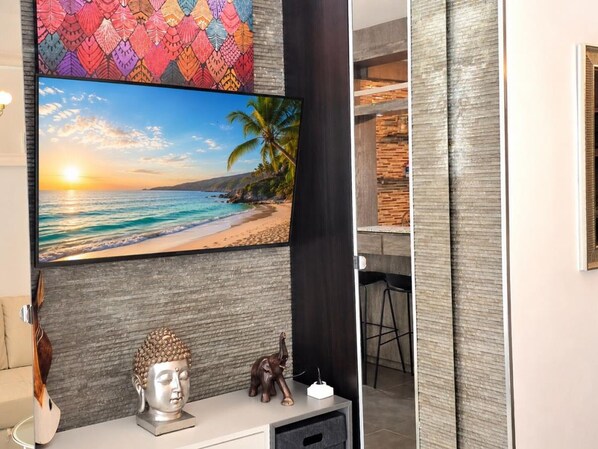 Une pièce avec un grand téléviseur diffusant une scène de plage, une statue de Bouddha et une petite figurine d’éléphant posée sur une console blanche.