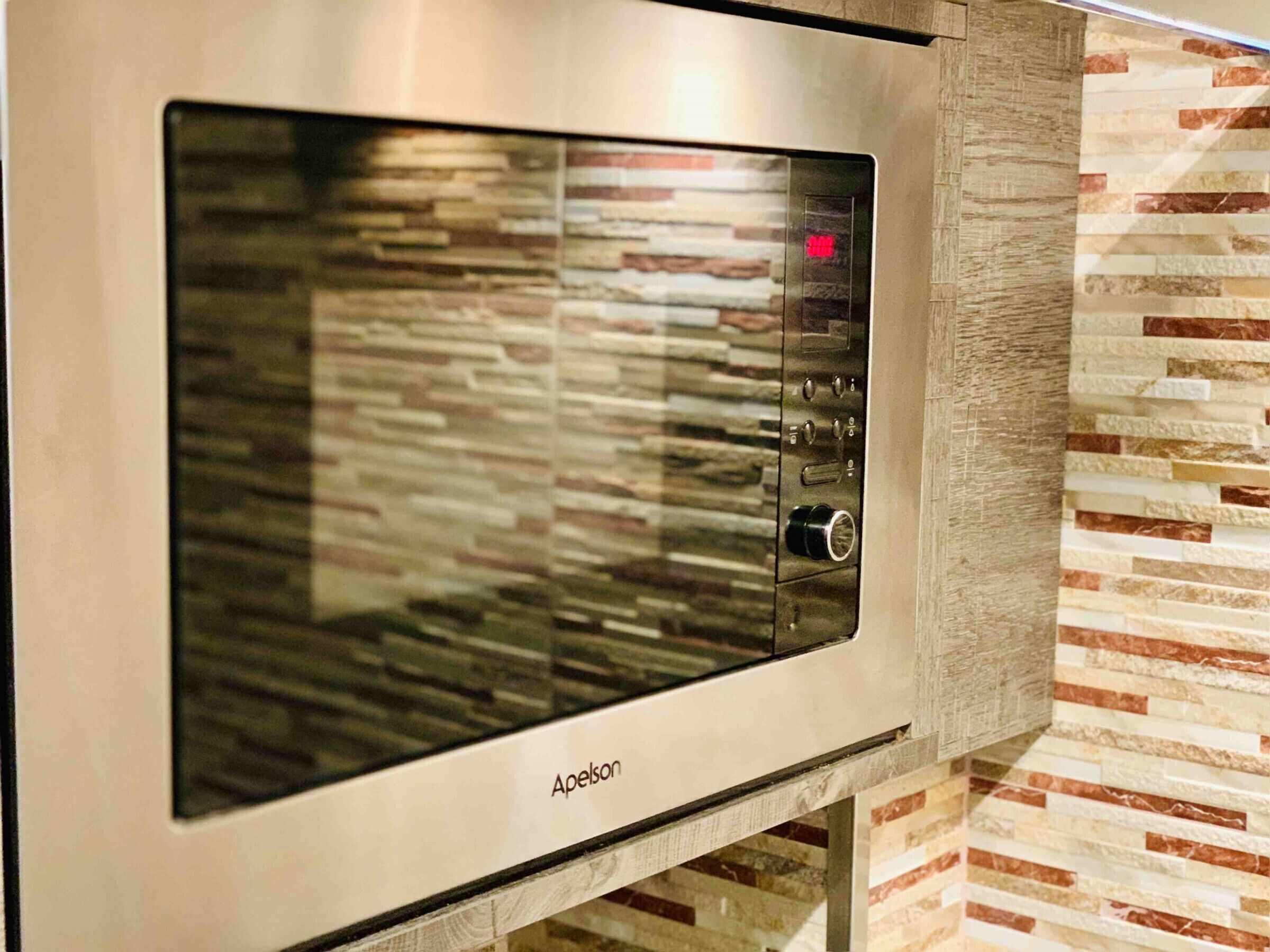 A stainless steel microwave oven with a digital display and control panel, mounted on a wall with a textured stone-like backsplash.