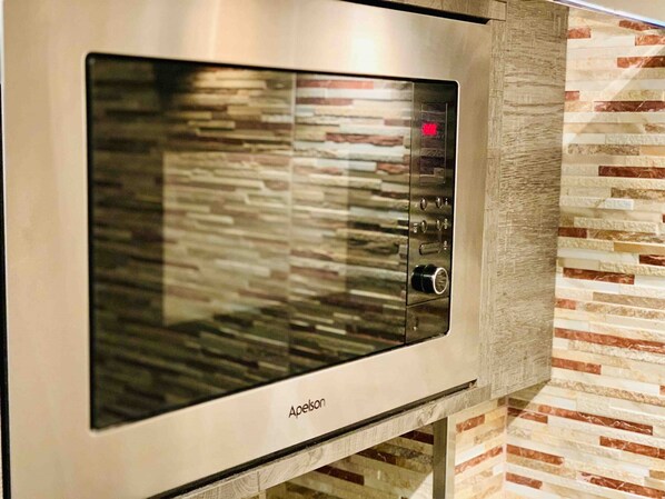 A stainless steel microwave oven with a digital display and control panel, mounted on a wall with a textured stone-like backsplash.