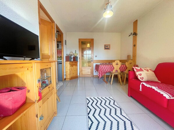 A compact living space with a television, wooden cabinets, a red sofa, and a dining area with a table and chairs.