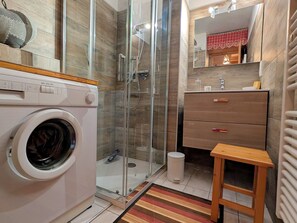 A compact bathroom with a washing machine, a glass shower enclosure, a wooden stool, and a vanity with drawers.