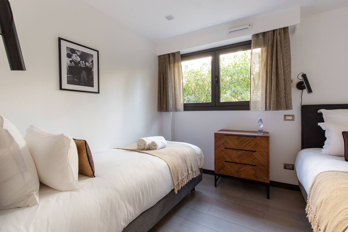 A bedroom with a bed, a nightstand, a dresser, a window with curtains, and a wall-mounted lamp.