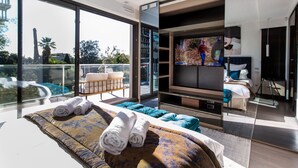 A modern bedroom with a large TV, a bed with rolled towels, a sofa with cushions, and a balcony with a view of the city. - Sweet Inn - Villa Alexandra (Cannes)