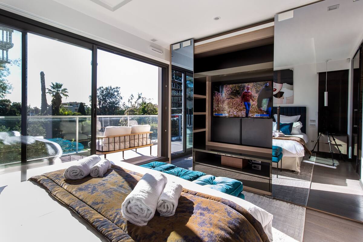 A modern bedroom with a large TV, a bed with rolled towels, a sofa with cushions, and a balcony with a view of the city.