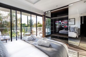 A modern bedroom with a large bed, a glass door leading to a balcony, and a view of the outdoors.